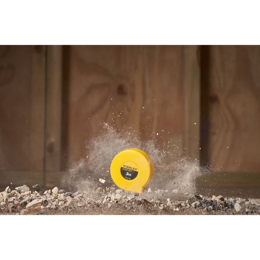 STANLEY 0-32-189 tape measure in yellow casing on rubble, impact dust in background, photography.