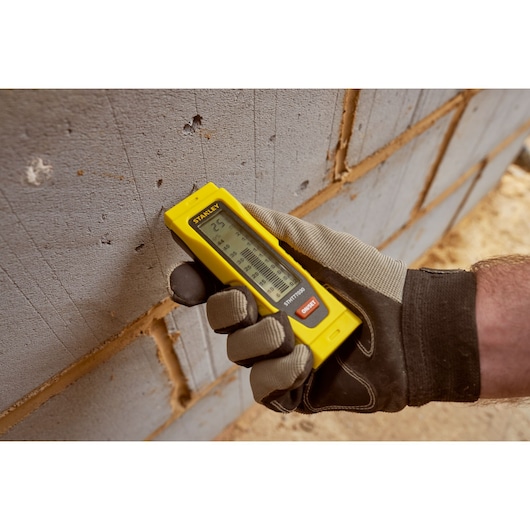 STANLEY 0-77-030 laser distance measurer held by gloved hand against brick wall, product in use.