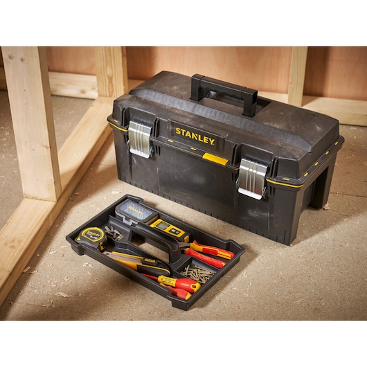 A black Stanley toolbox with metal latches and a handle is shown on the floor. The removable tray beside it contains various hand tools, including a screwdriver, utility knife, tape measure, spirit level, and some screws. The toolbox is placed in a workshop or construction area with wooden frames and sawdust visible.
