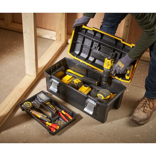 A person wearing gloves is opening a black and yellow STANLEY toolbox on a workshop floor. The toolbox contains various power tools and accessories, and a removable tray with hand tools and screws is placed beside it.