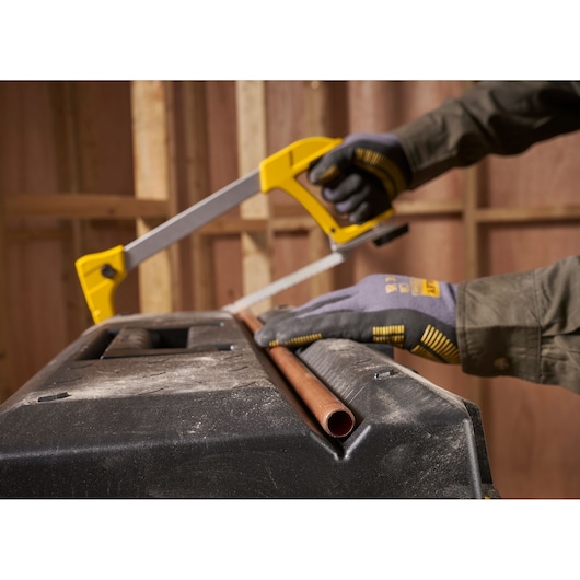 A person wearing work gloves uses a yellow and silver hacksaw to cut a copper pipe resting on a black workstation in a workshop setting.