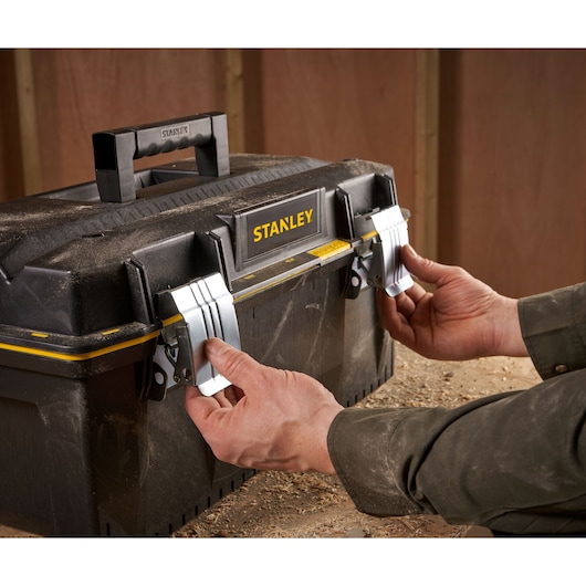 A person is opening the metal latches of a black Stanley toolbox with a yellow stripe, on a dusty workbench in a workshop.
