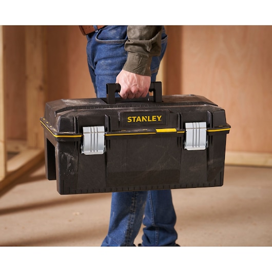 A person holding a black STANLEY toolbox with metal latches and a handle, shown in a workshop setting.