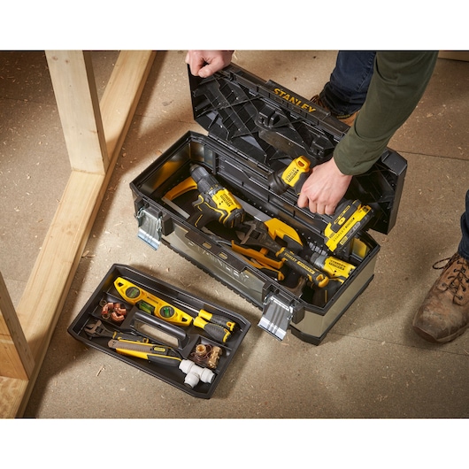 A STANLEY toolbox is open on the floor, showing various hand tools and power tools inside. The removable tray contains several small tools and accessories. A person is holding the lid, and construction materials are visible nearby.