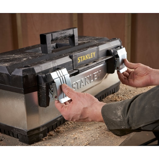 Hands opening the metal latches of a STANLEY toolbox placed on a workbench, with wood shavings around.