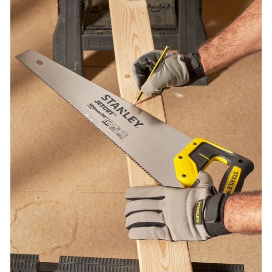 Stanley hand saw with yellow handle held on wood plank. Gloved hands mark the plank with a pencil for cutting.