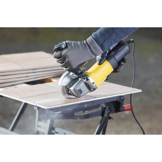 Photo of STANLEY FMEG210K angle grinder cutting tile, close-up with gloved hands and yellow handle.