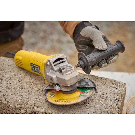 STANLEY FMEG615 angle grinder with yellow body, worker attaching side handle, outdoor concrete surface.