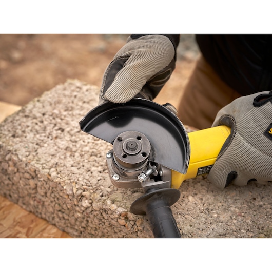 Photo of STANLEY FMEG615 angle grinder held by gloved hands on a concrete block, close-up view.