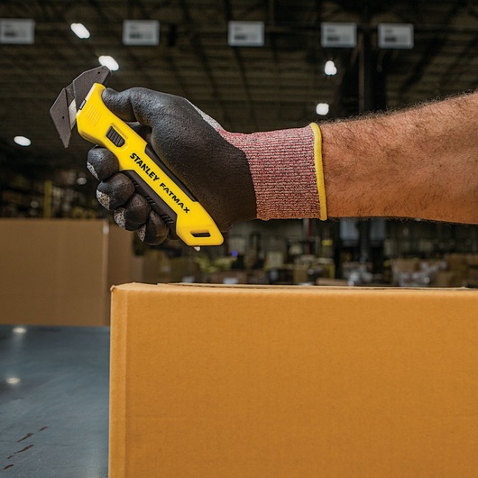 Photo of STANLEY FMHT10358 utility knife in gloved hand, cutting a cardboard box in a warehouse.
