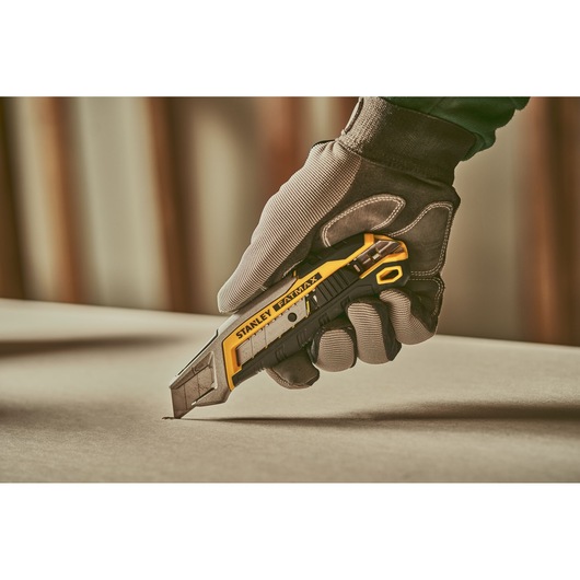 Stanley FMHT10594 utility knife held by gloved hand cutting material, photography close-up.