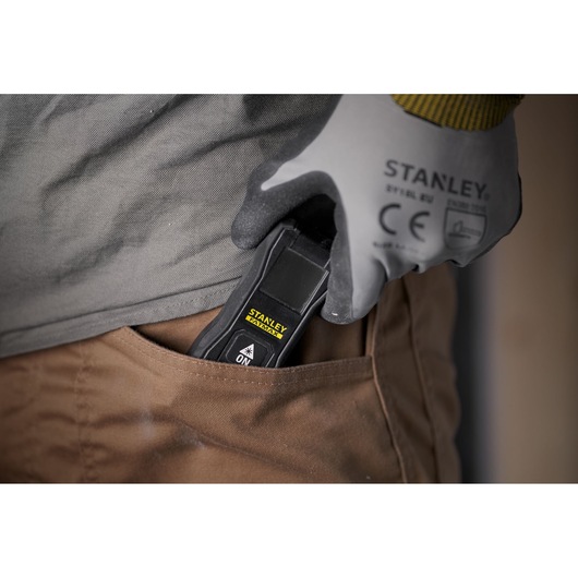 Photo of STANLEY FMHT77165-0 laser distance measurer in gloved hand, being placed in brown pants pocket.