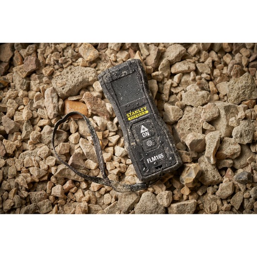 STANLEY FMHT77165-0 laser distance measurer with strap on rocky ground, covered in dust and water drops.