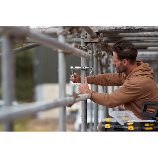STANLEY FMMT82678-0 ratchet in use on scaffolding, worker in brown hoodie, STANLEY toolbox nearby.
