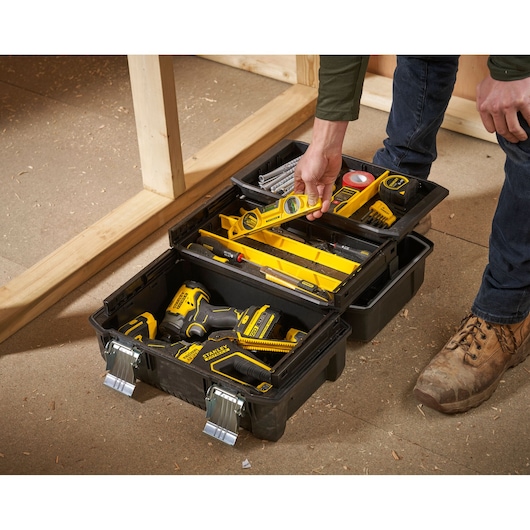 A person is reaching into an open STANLEY toolbox containing various tools, including a spirit level, a tape measure, and a drill. The toolbox is placed on a construction site floor.