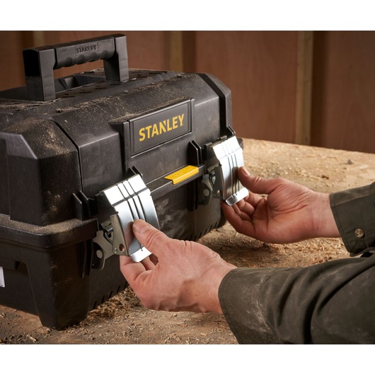 Hands opening the metal latches on a black STANLEY toolbox resting on a wooden surface.