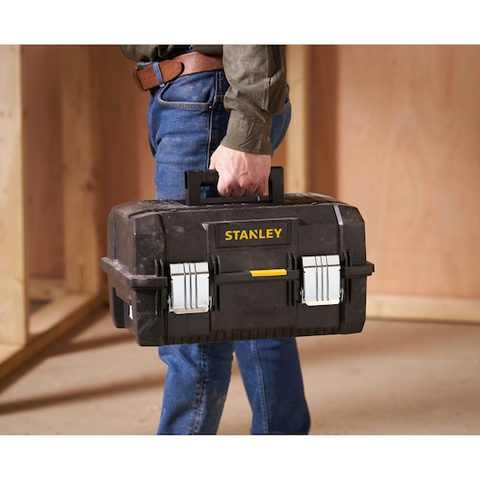 A person is holding a black STANLEY toolbox with metal latches and a yellow logo, standing in a workshop or construction area.