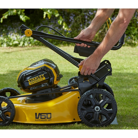 STANLEY SFMCMW2653B lawn mower side view, handle being adjusted, yellow and black design, outdoor grass setting.