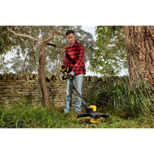 STANLEY SFMCSTB933B string trimmer in use outdoors, yellow and black, grass cutting near stone wall.