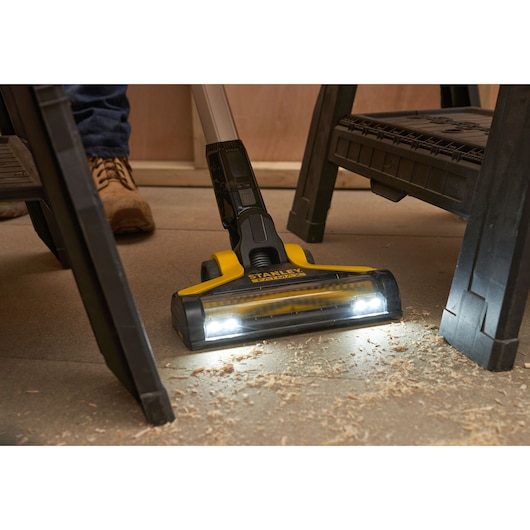 STANLEY SFMCVS001D1 cordless stick vacuum with LED lights cleaning wood shavings under a workbench, front angle.