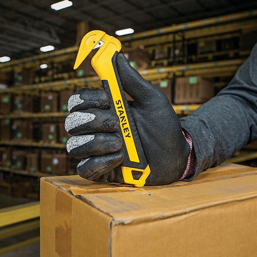 Stanley STHT10356 safety box cutter held by a gloved hand opening a cardboard box in a warehouse.