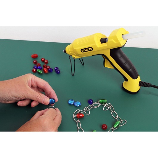 Stanley STHT6-70417 yellow glue gun for crafts shown with beads and jewelry chain on green surface.