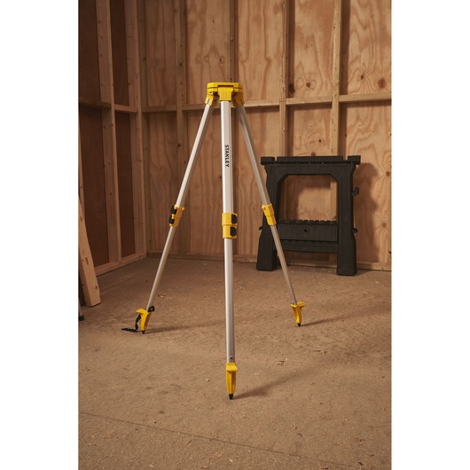 STANLEY STHT77625-1 aluminium tripod with yellow accents, standing in a wooden workshop, photography side angle.
