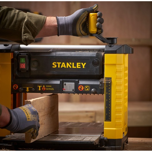 STANLEY STP18-B2 wood planer in use, close-up side angle, yellow and black, gloved hands adjusting controls.