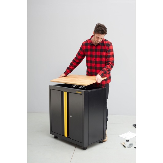 STANLEY STST97595-1 storage cabinet with black body, yellow stripe, shown in assembly with wood top being placed.